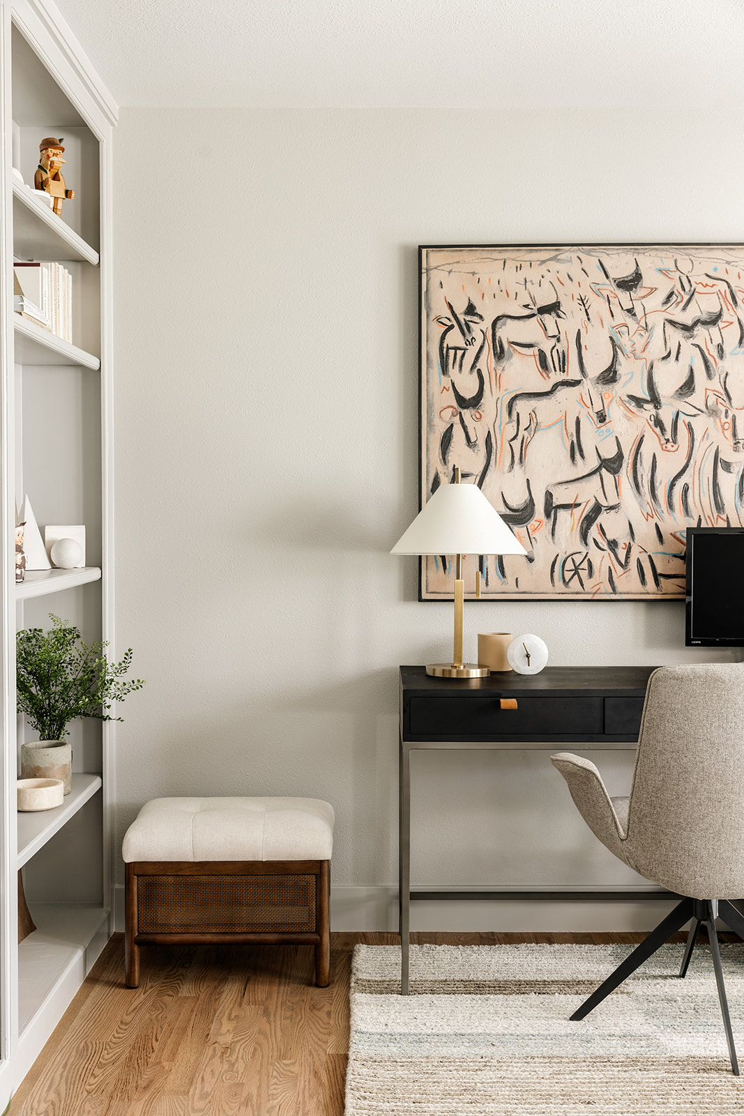Modern rustic home office featuring a black metal desk, neutral upholstered office chair, abstract art by Kader Boly, brass task lamp, and built-in shelving with organic styling.